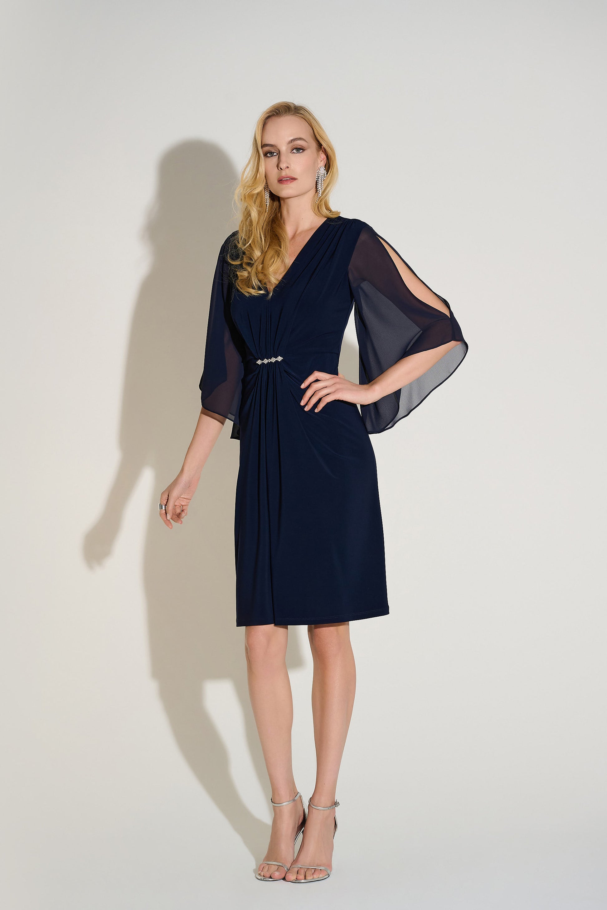 Hannahs Of Erin| Erin Ontario|Joseph Ribkoff Signature silky knit and chiffon sheath dress in midnight blue, featuring a V-neckline, three-quarter chiffon bell sleeves, delicate front draping, and a rhinestone ornament at the waist. Falls just below the knee and fully lined for a smooth, elegant fit, ideal for special occasions.
