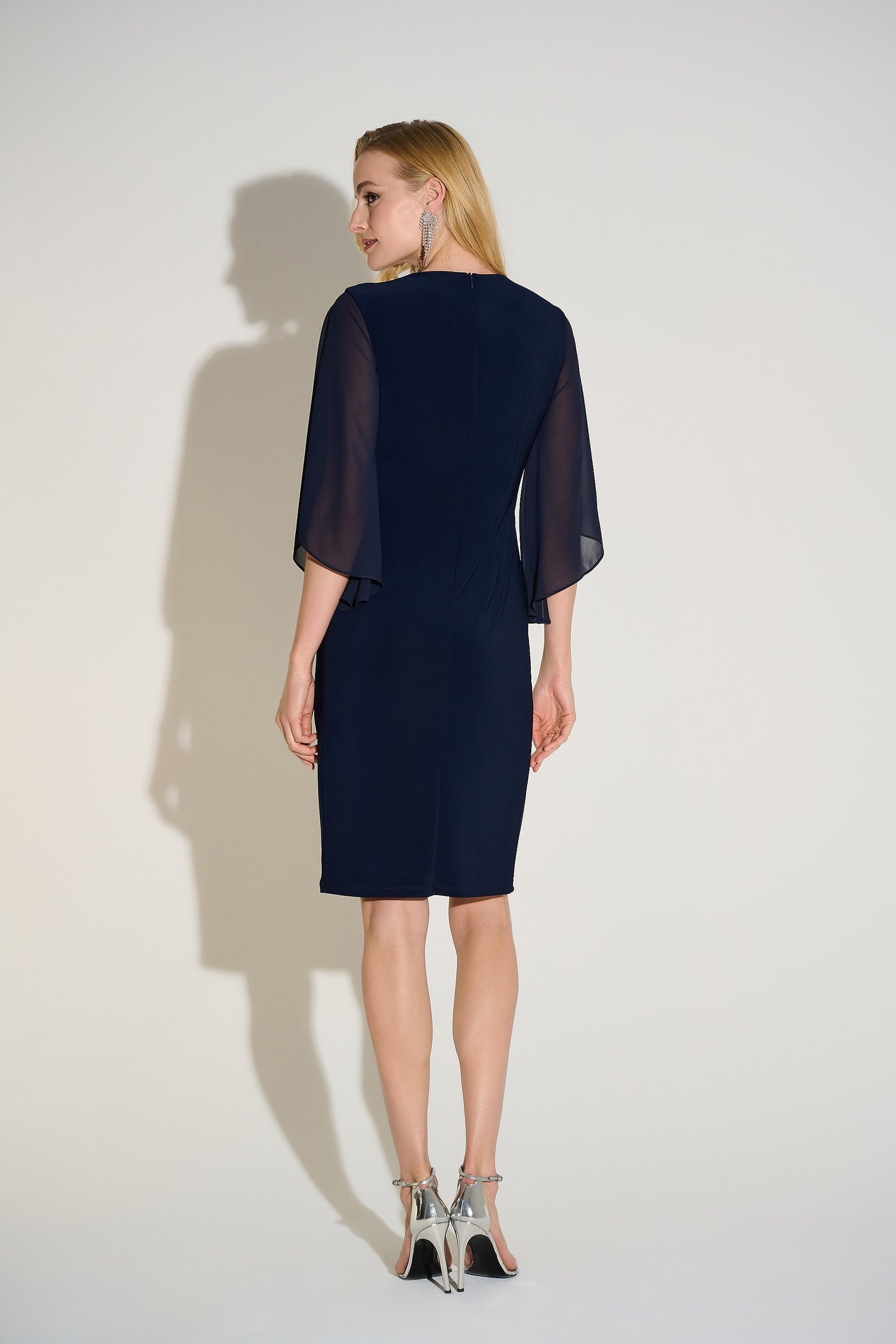 Hannahs Of Erin| Erin Ontario|Joseph Ribkoff Signature silky knit and chiffon sheath dress in midnight blue, featuring a V-neckline, three-quarter chiffon bell sleeves, delicate front draping, and a rhinestone ornament at the waist. Falls just below the knee and fully lined for a smooth, elegant fit, ideal for special occasions.
