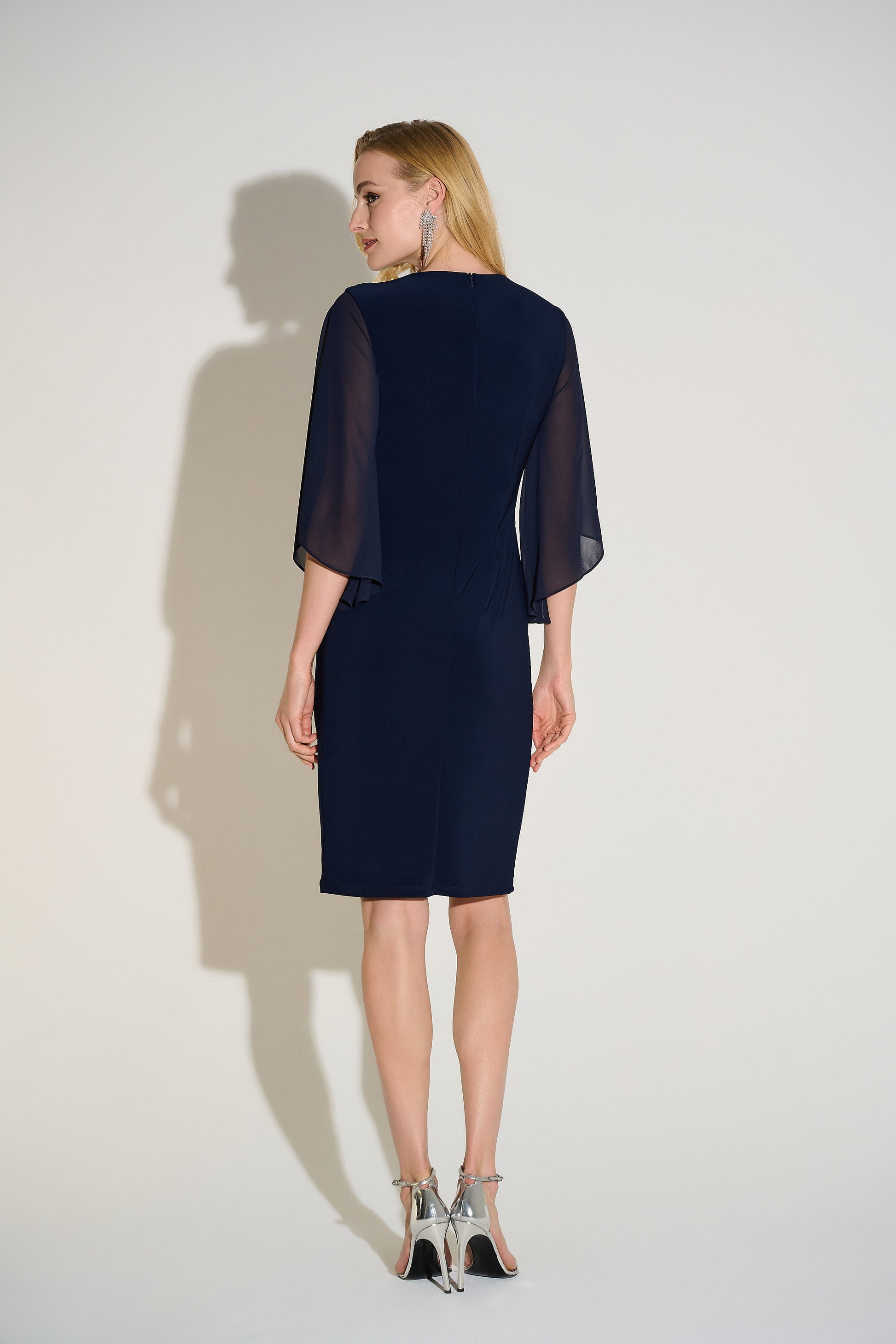 Hannahs Of Erin| Erin Ontario|Joseph Ribkoff Signature silky knit and chiffon sheath dress in midnight blue, featuring a V-neckline, three-quarter chiffon bell sleeves, delicate front draping, and a rhinestone ornament at the waist. Falls just below the knee and fully lined for a smooth, elegant fit, ideal for special occasions.
